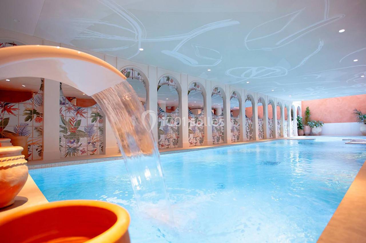 Piscine intérieure avec jets d'eau, décorée de fresques colorées et entourée d'arches lumineuses.