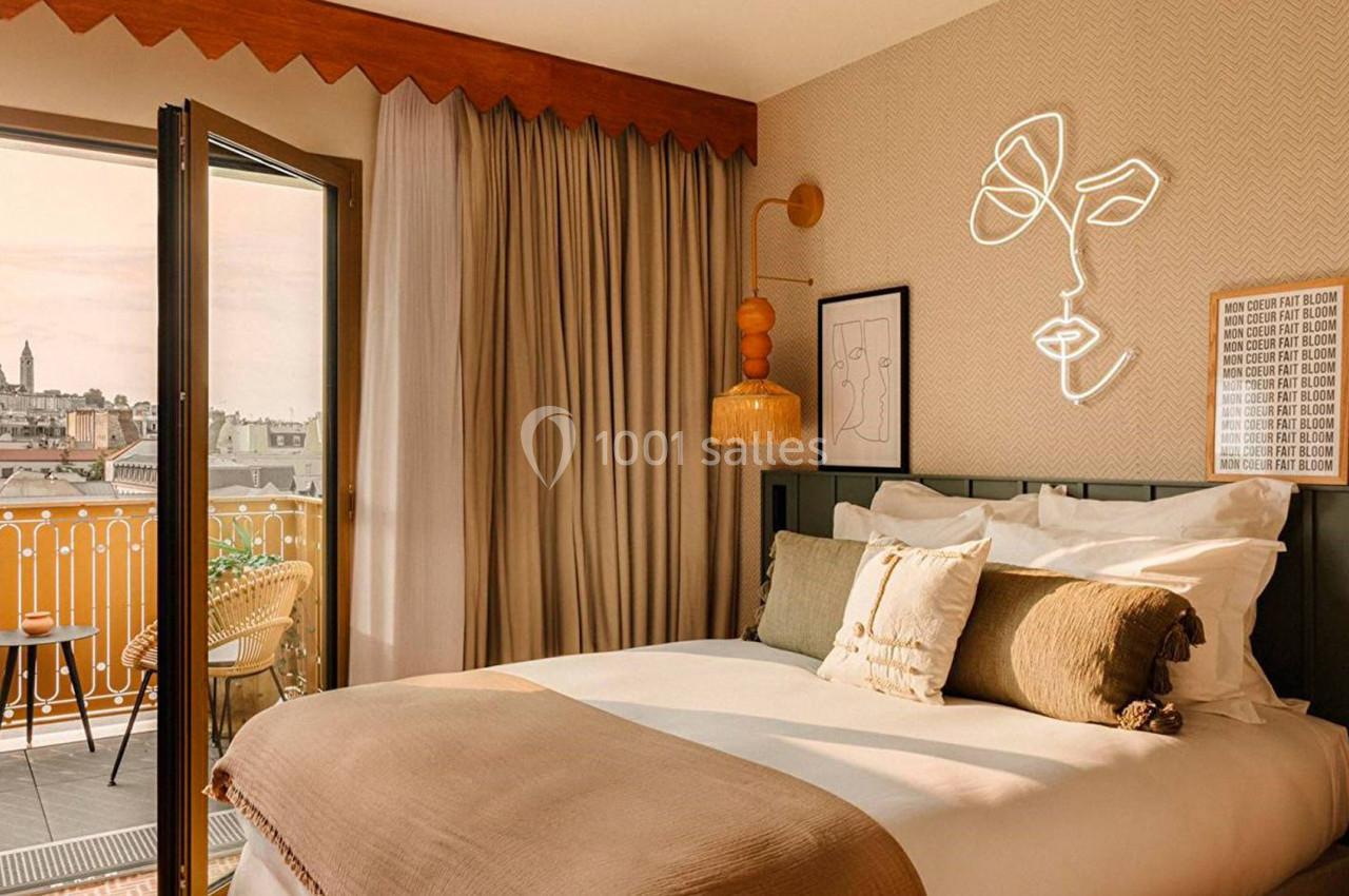 Chambre lumineuse avec lit double, décoration moderne, balcon ouvert sur une vue urbaine et éclairage chaleureux.