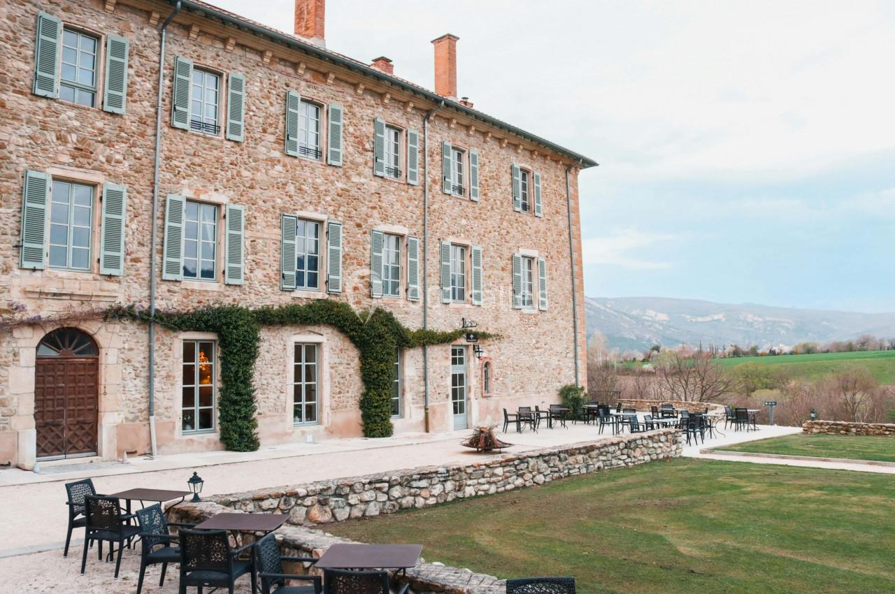 Façade en pierre d'un bâtiment ancien avec terrasse aménagée, entourée de verdure et vue sur la campagne.