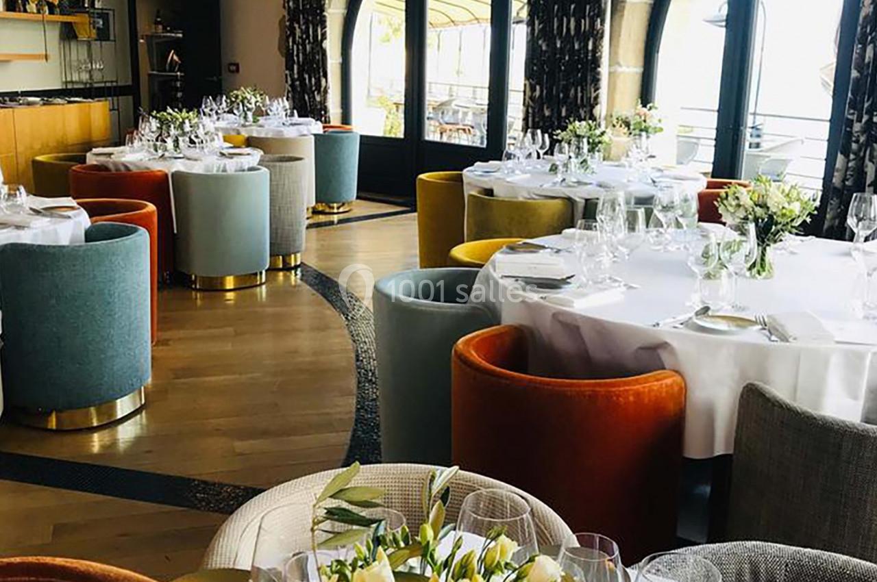 Salle de restaurant élégante avec tables rondes dressées, chaises colorées et grandes fenêtres donnant sur l'extérieur.
