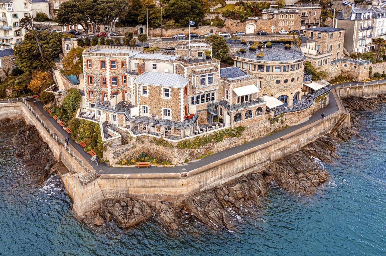 Vue aérienne d'un bâtiment en pierre sur une falaise entourée de mer, avec des arbres et des maisons en arrière-plan.
