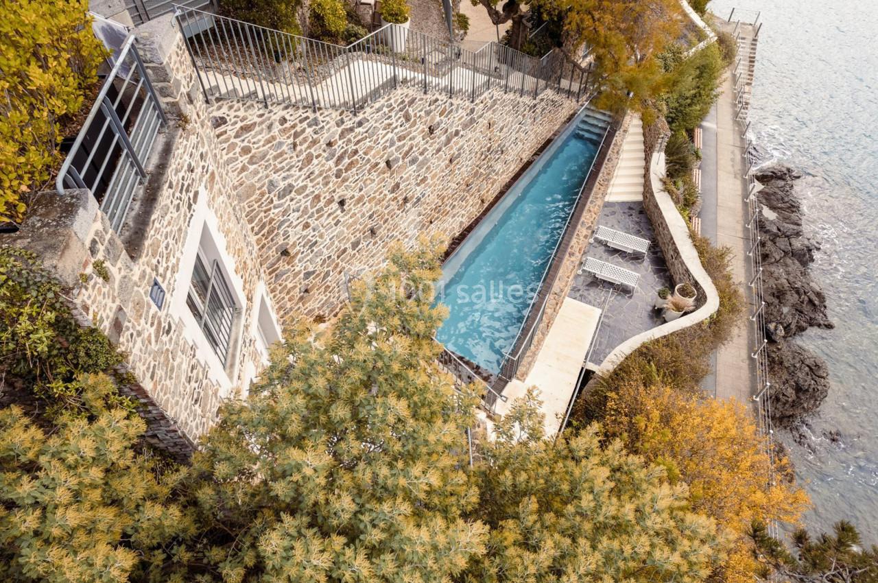 Piscine étroite entourée de murs en pierre, située en bord de mer avec végétation et chemin adjacent.