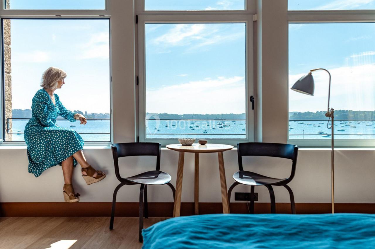 Une femme assise sur le rebord d'une fenêtre contemple une vue sur la mer, dans une pièce lumineuse et moderne.
