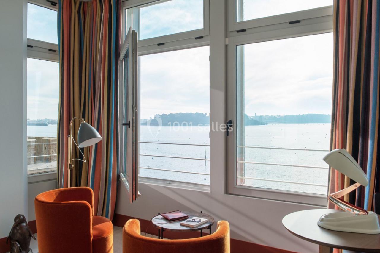 Vue sur la mer depuis une pièce lumineuse avec fauteuils orange, lampes modernes et fenêtres ouvertes.
