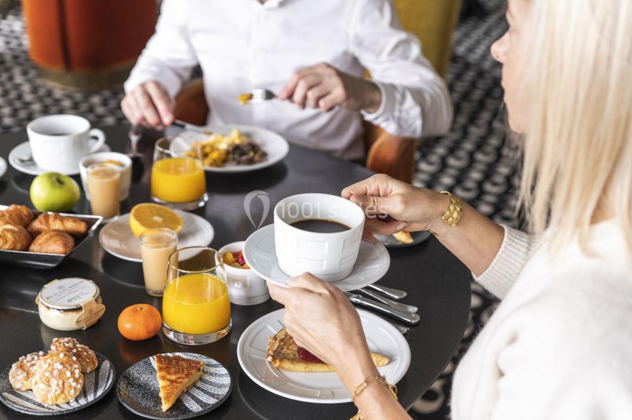 Une femme tient une tasse de café à table, entourée de viennoiseries, fruits et jus, lors d'un petit-déjeuner.