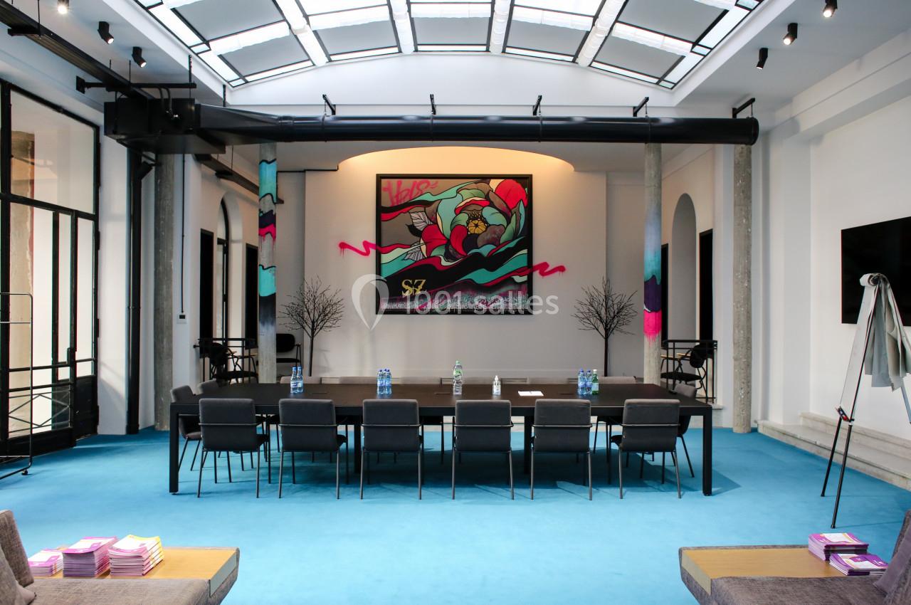 Salle de réunion moderne avec table, chaises, sol bleu, et une grande œuvre d'art colorée accrochée au mur.