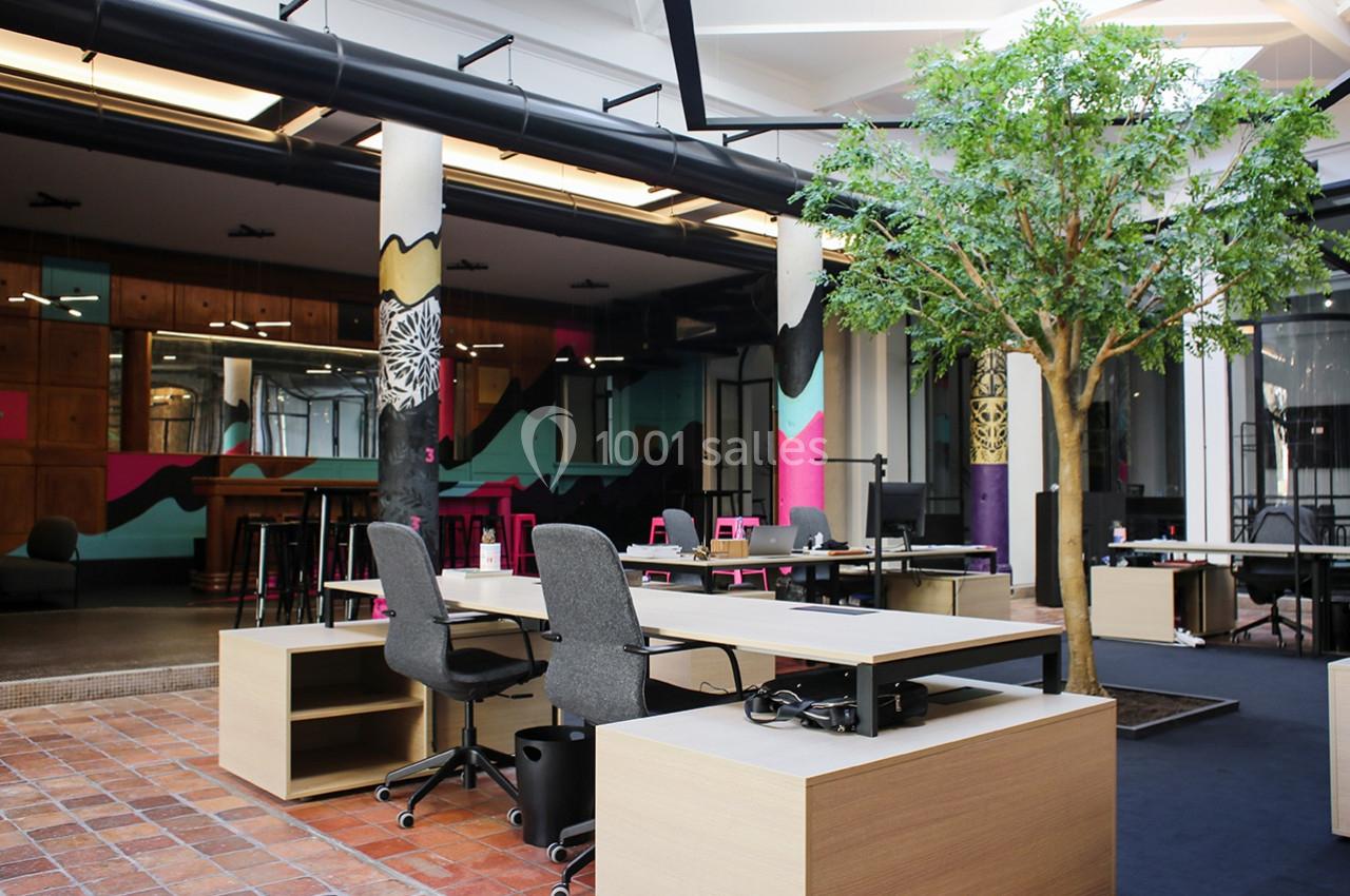 Espace de travail moderne avec bureaux partagés, sièges ergonomiques, arbre décoratif et décor coloré en arrière-plan.