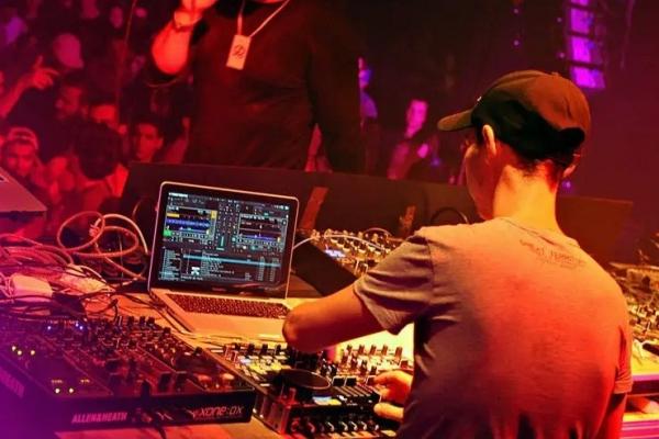 Un DJ mixe de la musique sur une table de mixage avec un ordinateur portable, devant un public dans une ambiance lumineuse.