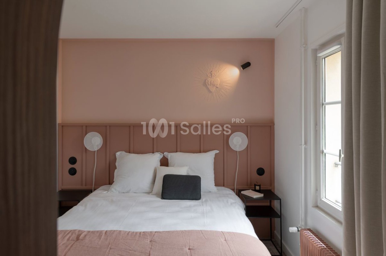 Chambre lumineuse avec lit double, tête de lit rose, lampes murales et fenêtre laissant entrer la lumière naturelle.