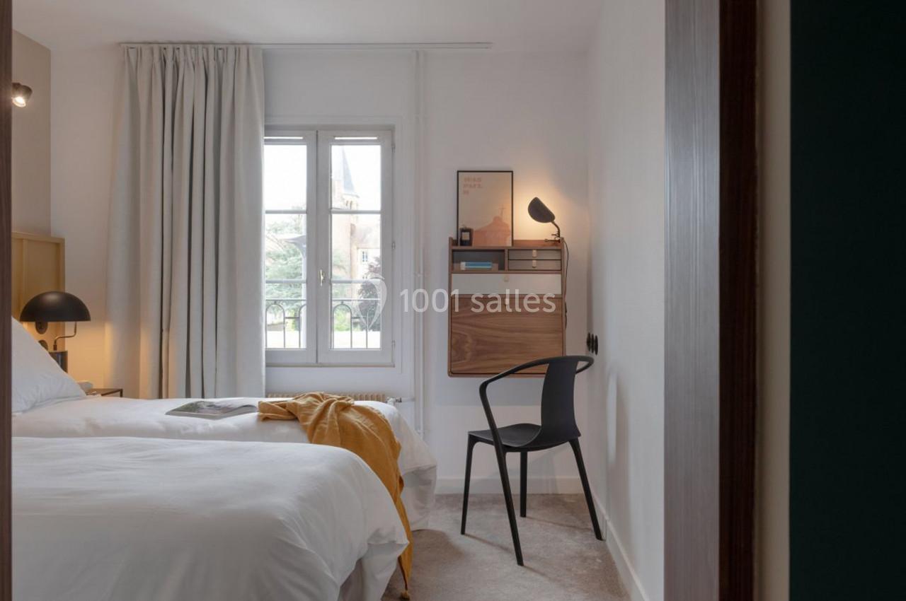 Chambre lumineuse avec lit double, bureau mural en bois, chaise noire et fenêtre donnant sur un paysage extérieur.