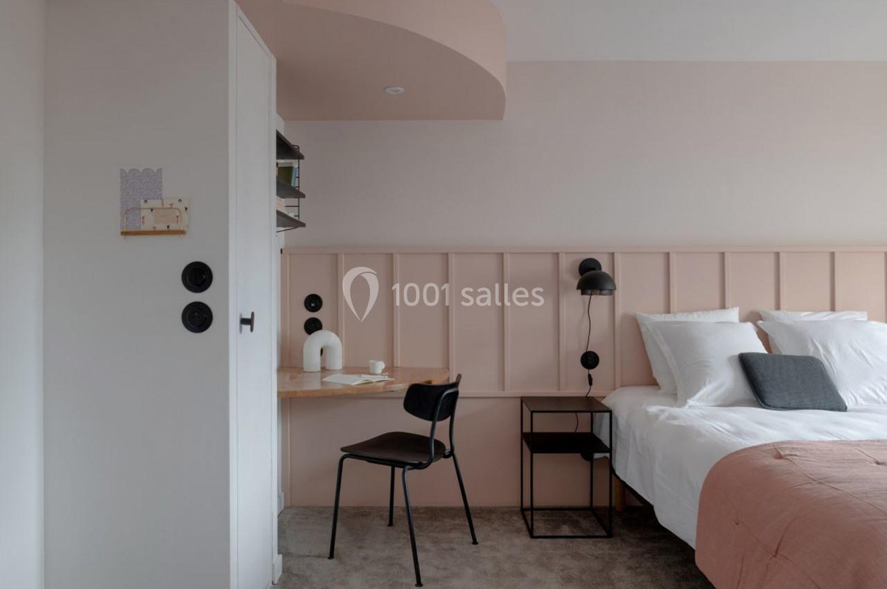 Chambre moderne avec lit double, mur rose, bureau en bois clair, chaise noire et éclairage mural.