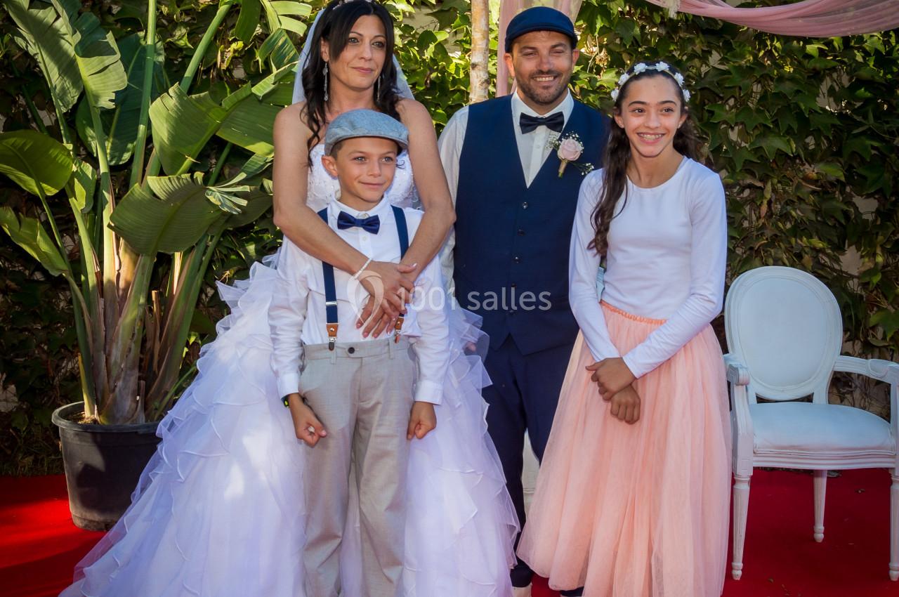 Un couple en tenue de mariage pose avec deux enfants sur un tapis rouge, devant un décor végétal et floral.