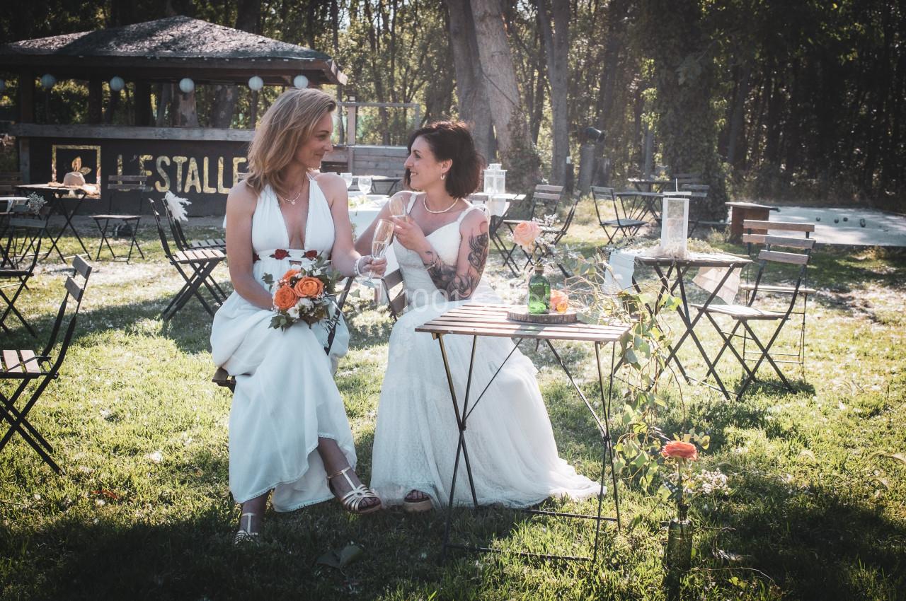 Deux femmes en robes de mariée assises dans un jardin, souriantes, tenant des fleurs et entourées de chaises.