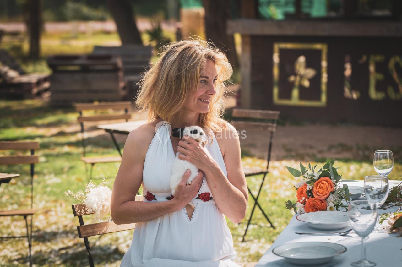 Une femme en robe blanche tient un lapin dans ses bras, assise à une table décorée de fleurs en extérieur.