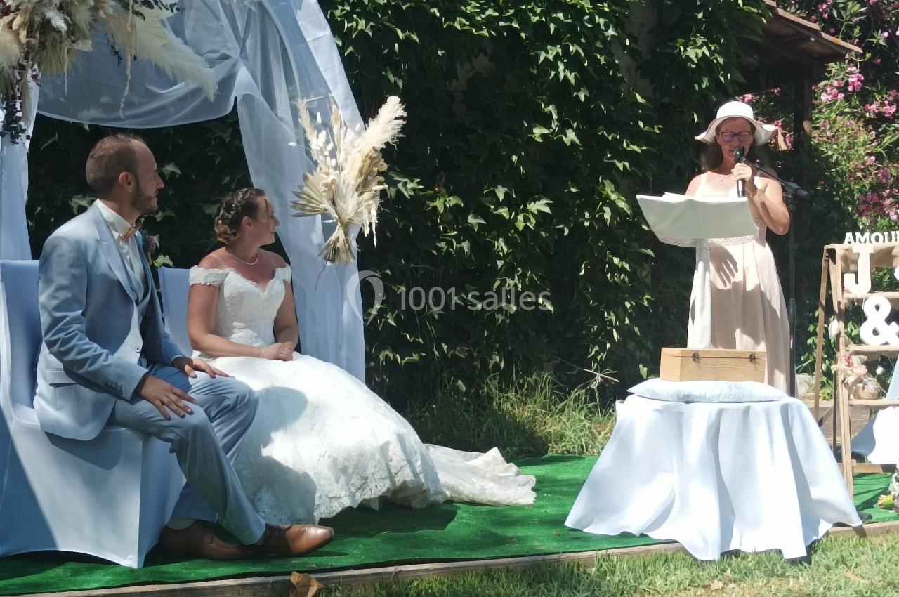 Un couple assis sous une arche décorée lors d'une cérémonie en plein air, avec une personne lisant un discours.