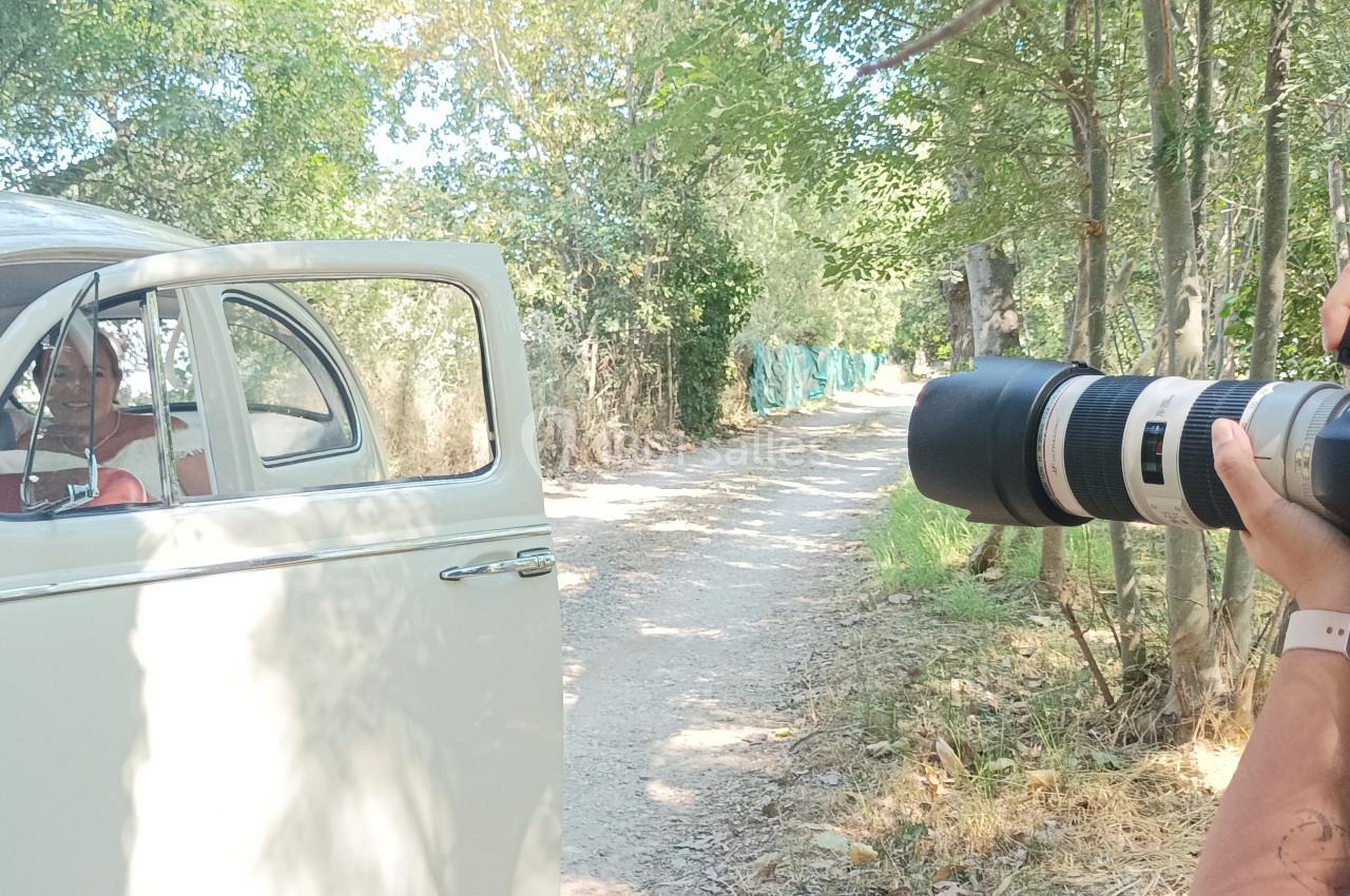 Un photographe capture un couple assis dans une voiture ancienne, garée sur un chemin bordé d'arbres.