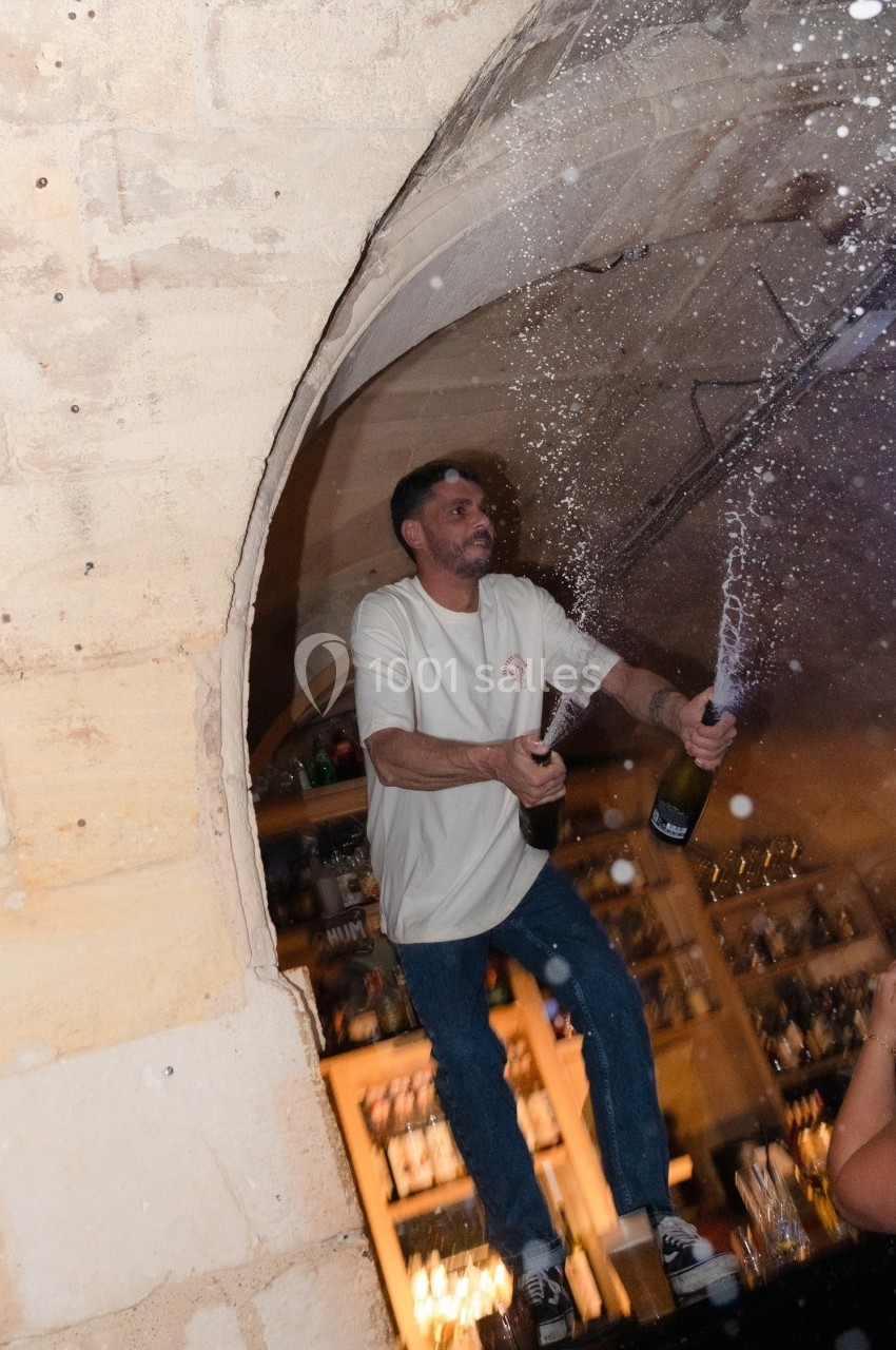 Un homme debout sur un bar fait jaillir du champagne de deux bouteilles dans un lieu à l'ambiance festive.