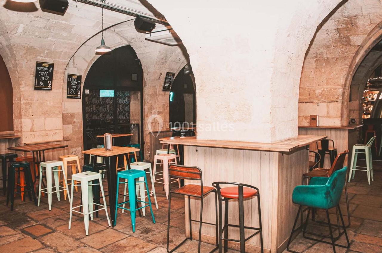 Salle voûtée en pierre avec des tables hautes, des tabourets colorés et un bar en bois clair.
