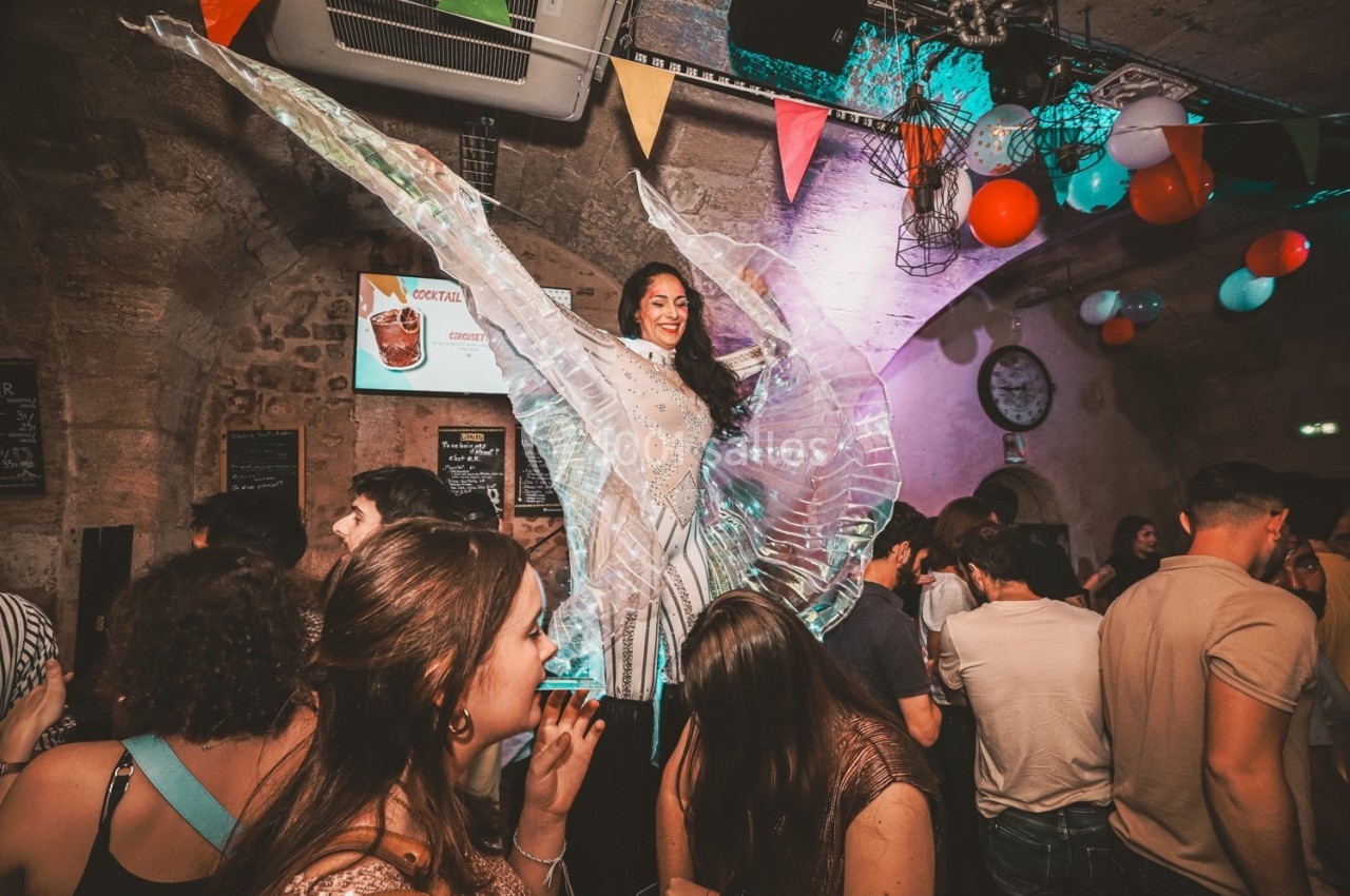 Une performeuse en costume lumineux danse au-dessus d'une foule dans un bar décoré de guirlandes et ballons colorés.