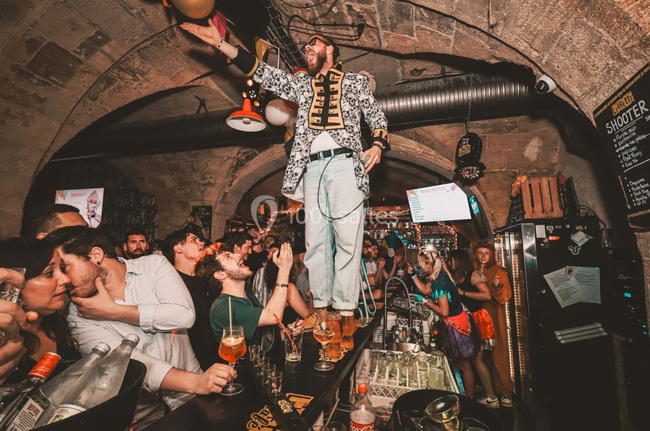 Un homme debout sur un bar animé, entouré de clients dans une ambiance festive et éclairage tamisé.