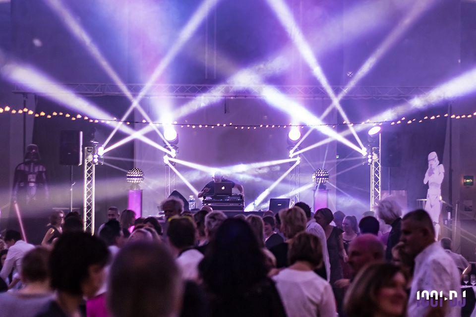 Foule dansante sous des jeux de lumière colorés, avec un DJ sur scène et une ambiance festive en intérieur.