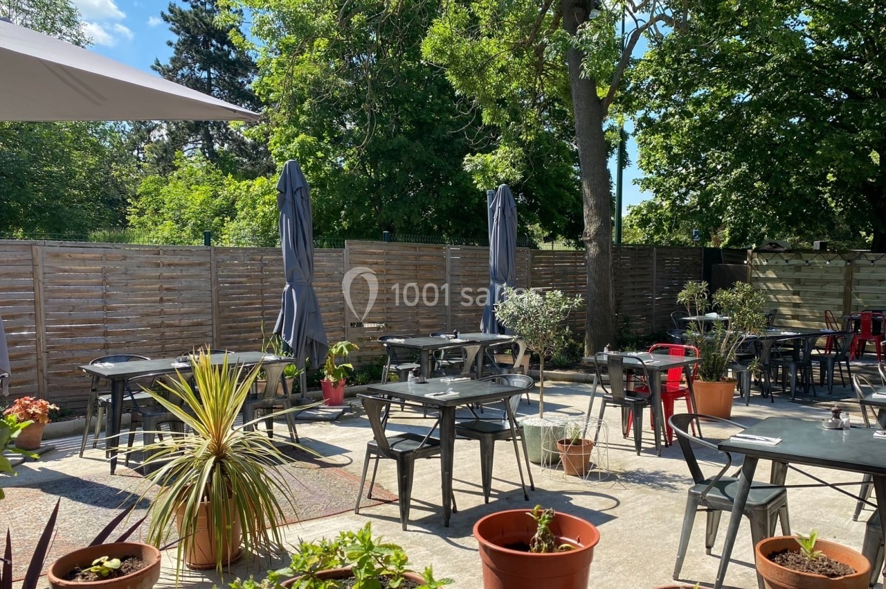 Terrasse extérieure avec tables, chaises, parasols et plantes en pots, entourée de végétation et d'une clôture en bois.