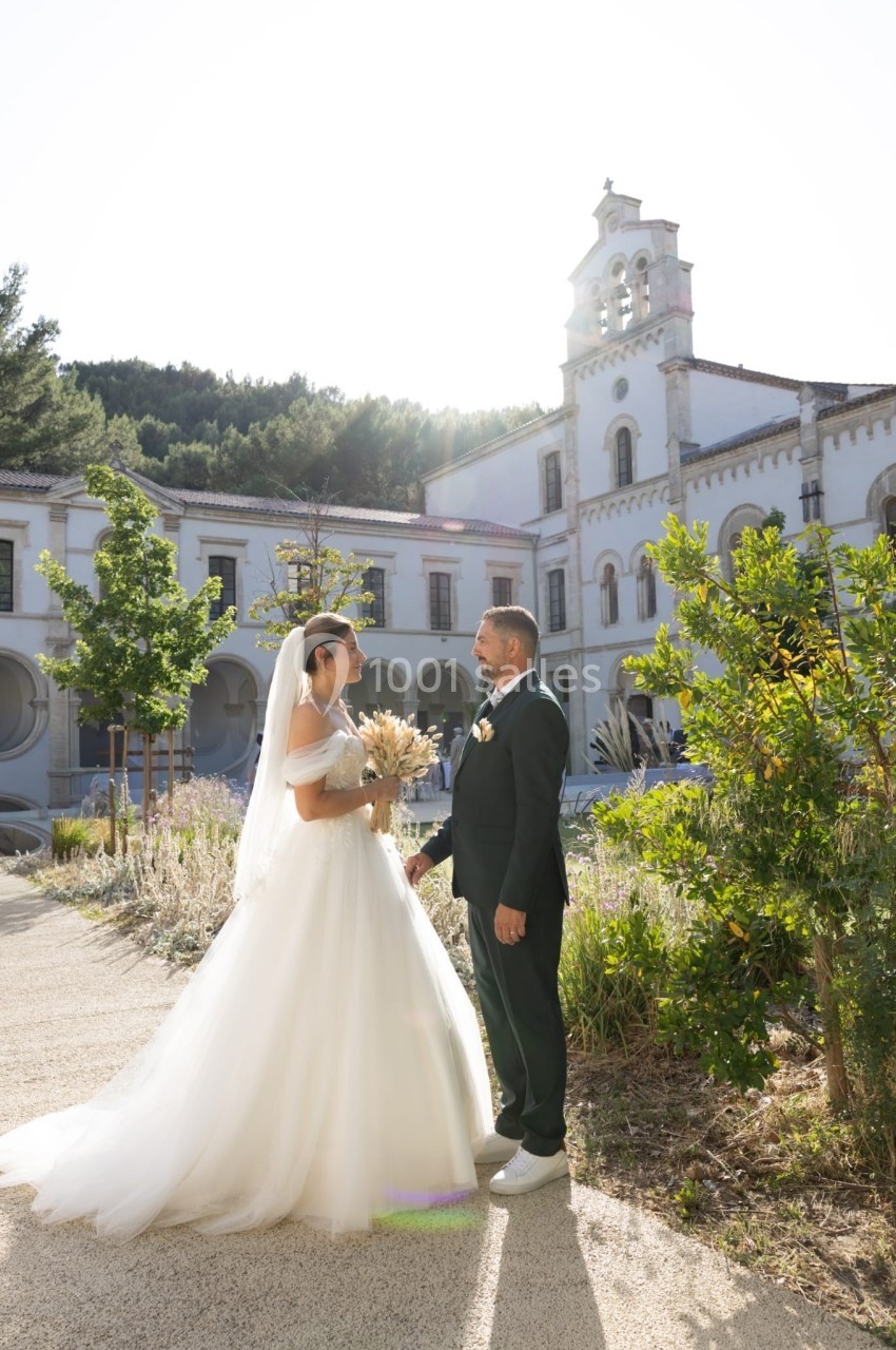 Un couple de mariés se tient face à face dans un jardin ensoleillé devant un bâtiment religieux.