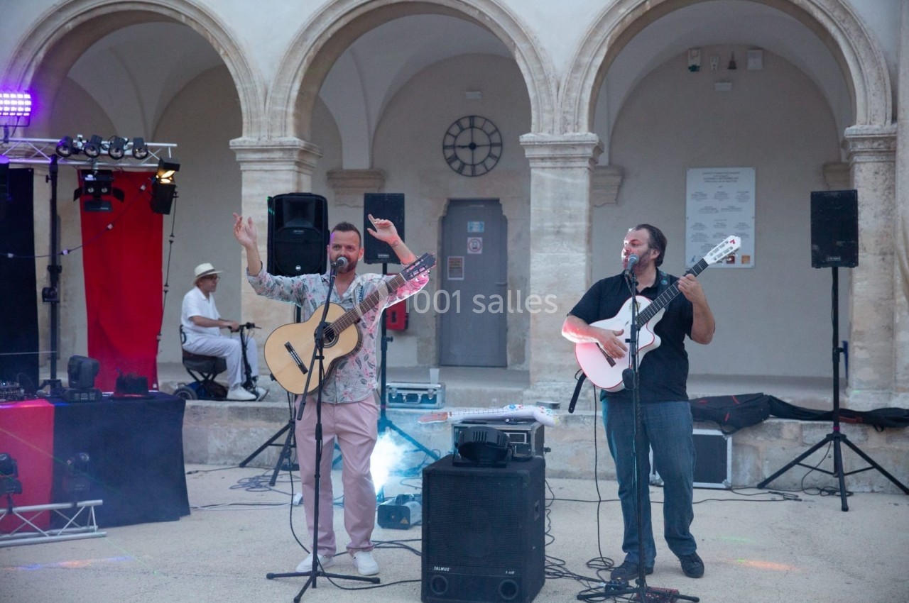 Deux musiciens jouent de la guitare et chantent sur une scène en plein air, devant des arches en pierre.