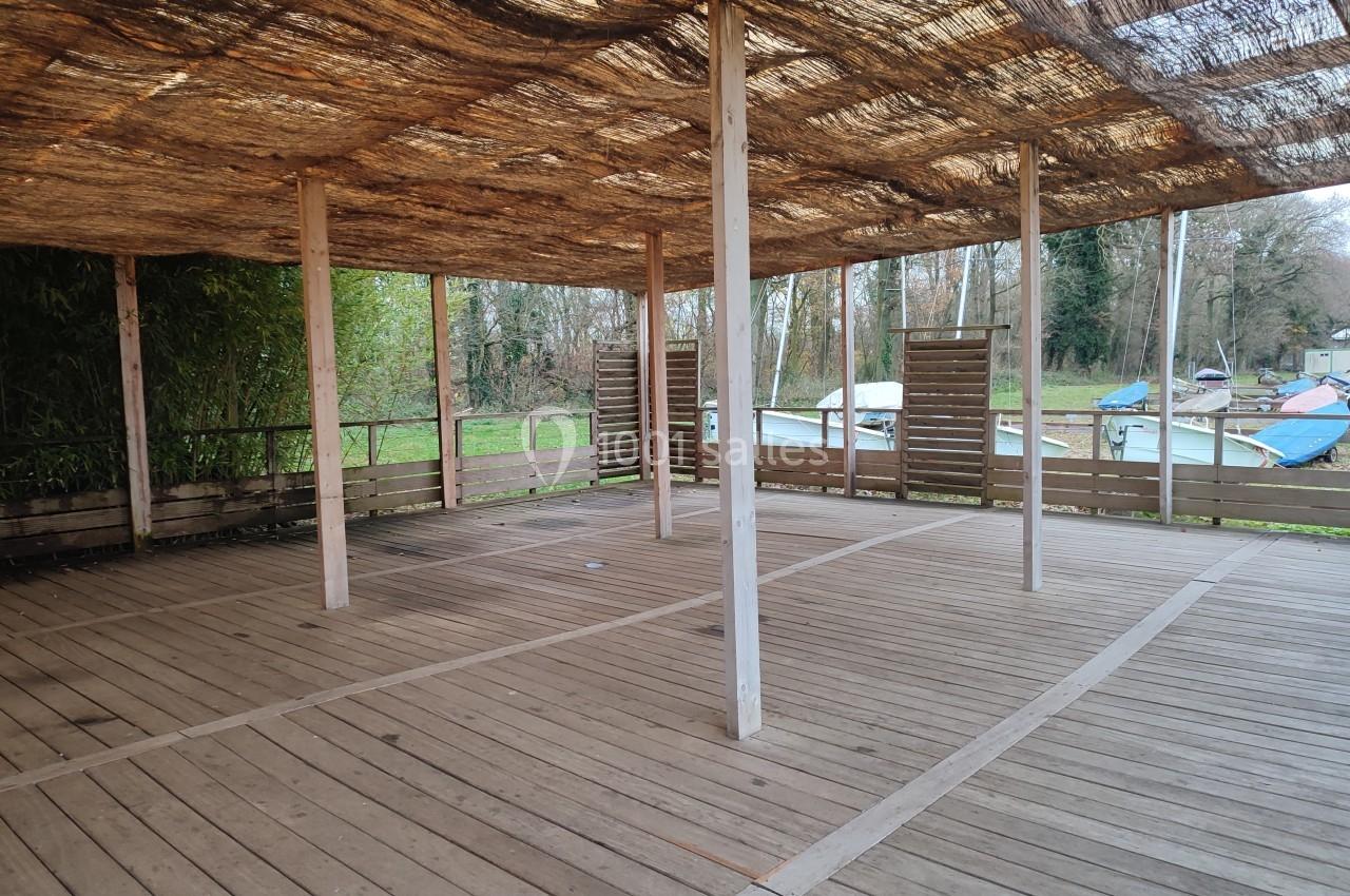 Structure en bois avec toit en canisses, ouverte sur les côtés, située près d'un espace naturel avec des bateaux visibles.