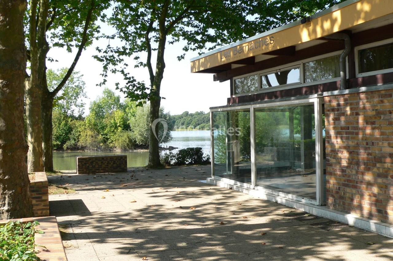 Bâtiment en briques avec de grandes baies vitrées, entouré d'arbres, près d'un plan d'eau sous un ciel dégagé.