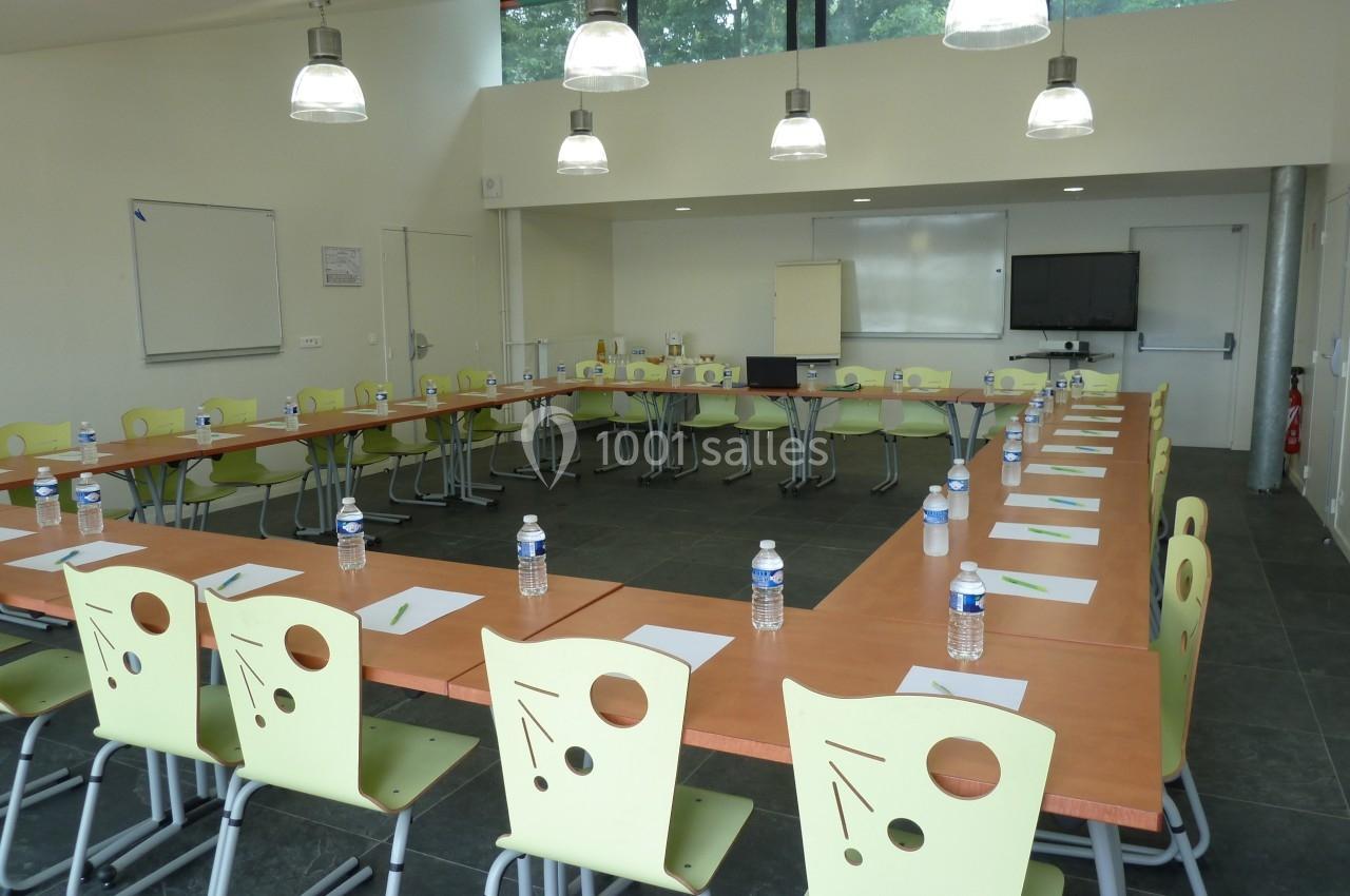 Salle de réunion lumineuse avec tables disposées en U, chaises vertes, bouteilles d'eau et matériel de présentation.