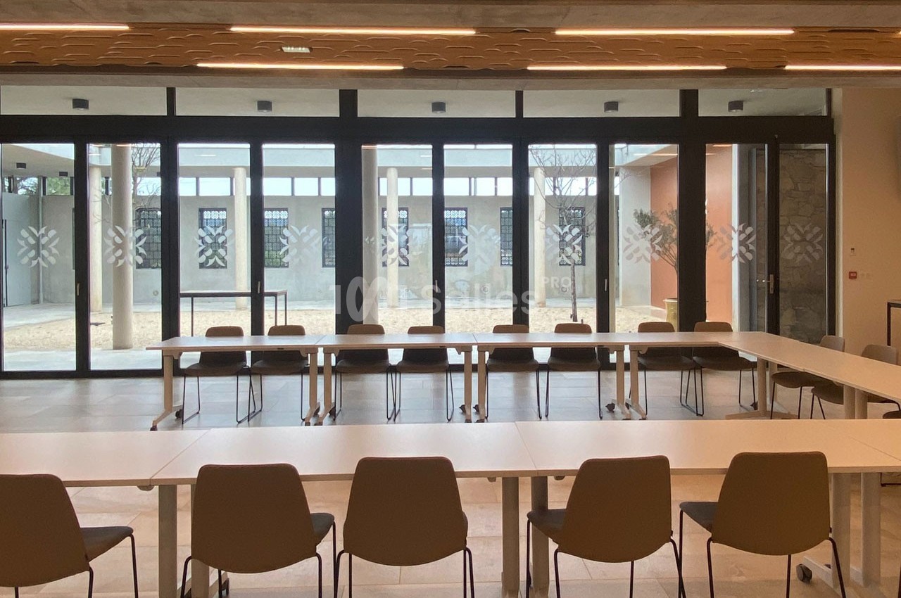 Salle de réunion lumineuse avec des tables disposées en U, des chaises et une grande baie vitrée donnant sur une cour.
