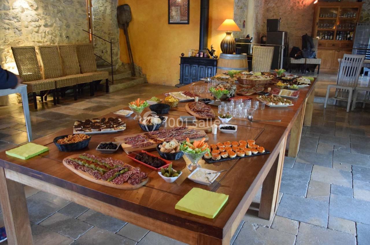 Table en bois garnie de plats variés, incluant charcuterie, légumes, desserts et apéritifs, dans une salle rustique.