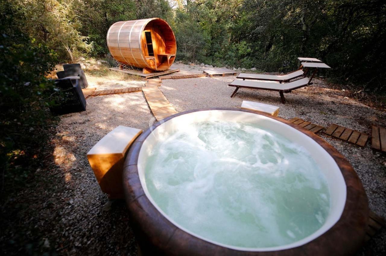 Bain à remous extérieur entouré de chaises longues et d'un sauna en bois dans un cadre naturel boisé.