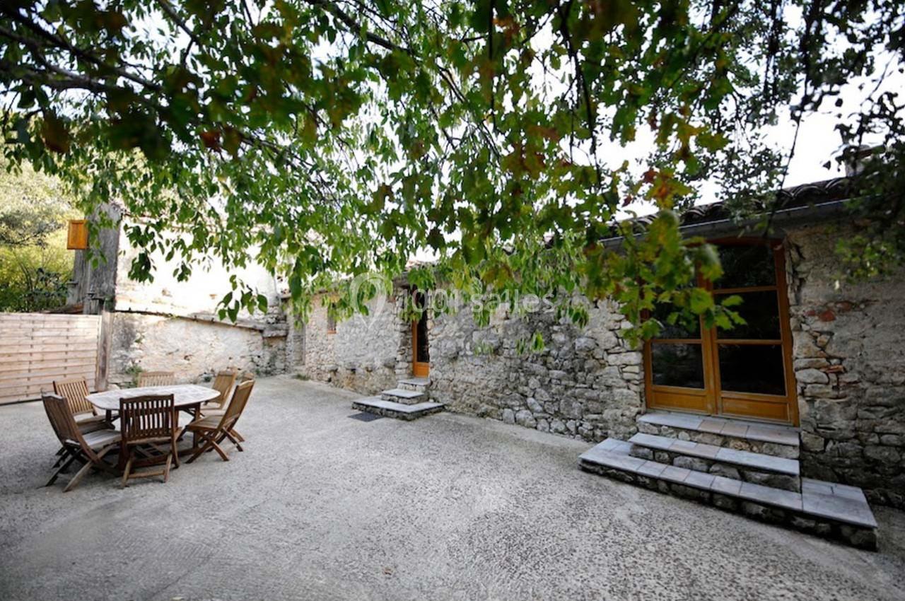 Cour extérieure avec table et chaises en bois, entourée de murs en pierre et partiellement ombragée par des arbres.