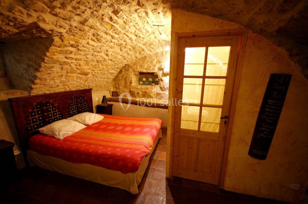 Chambre avec murs en pierre apparente, lit double avec couvre-lit rouge et porte vitrée donnant sur une pièce éclairée.