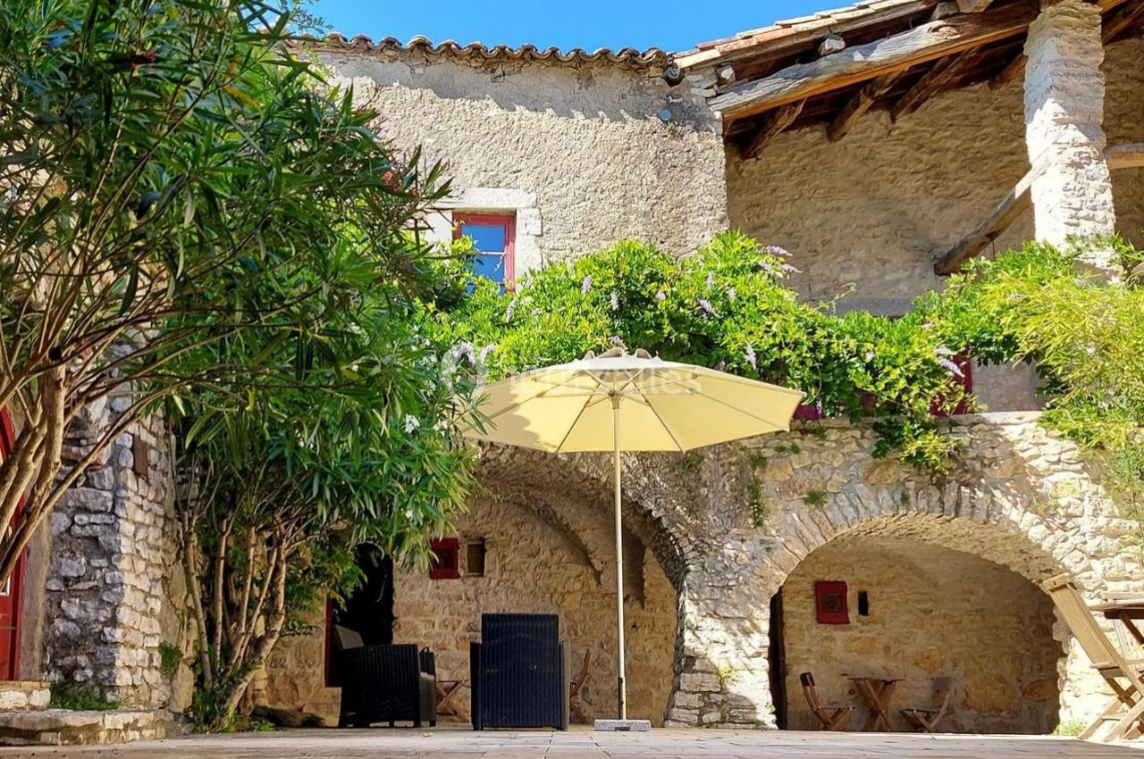 Cour ensoleillée avec murs en pierre, arches fleuries, mobilier d'extérieur et un grand parasol beige.