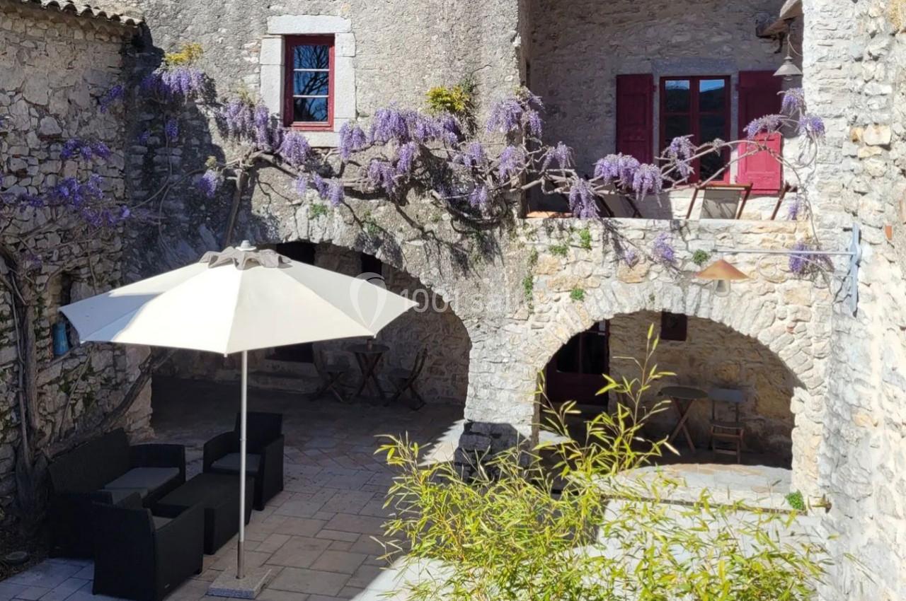 Cour en pierre avec mobilier de jardin, ombragée par un parasol blanc, et ornée de glycines en fleurs sur les arches.