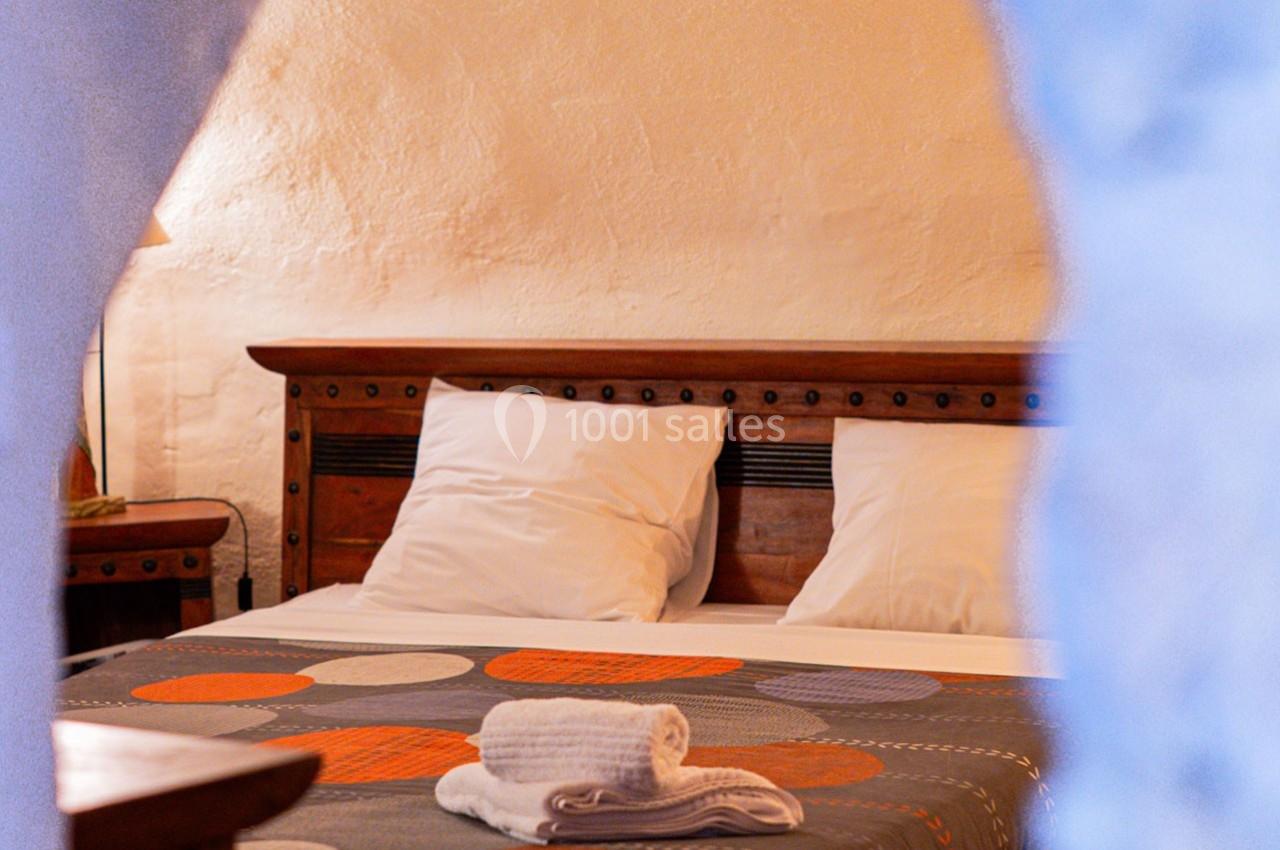 Lit double avec tête de lit en bois, draps blancs, couverture à motifs et serviettes pliées dans une chambre aux murs clairs.