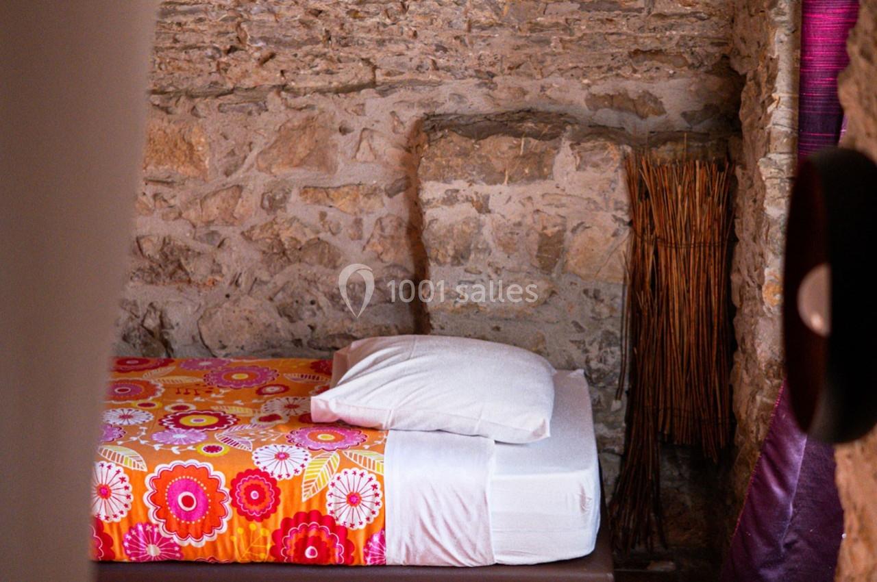 Lit simple avec une housse colorée à motifs floraux, adossé à un mur en pierre apparente dans une pièce rustique.