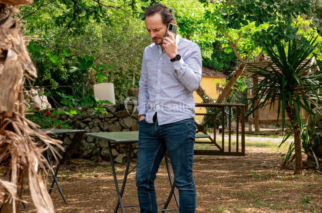 Homme debout dans un jardin, tenant un téléphone à l'oreille, entouré de végétation et de mobilier extérieur.