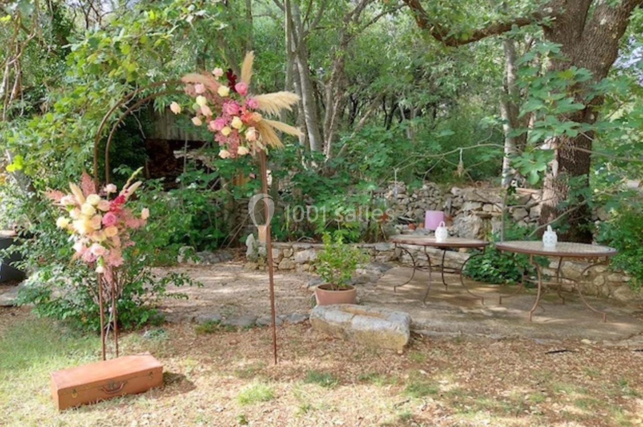 Arche florale décorée de fleurs pastel dans un jardin boisé, avec tables et pierres en arrière-plan.