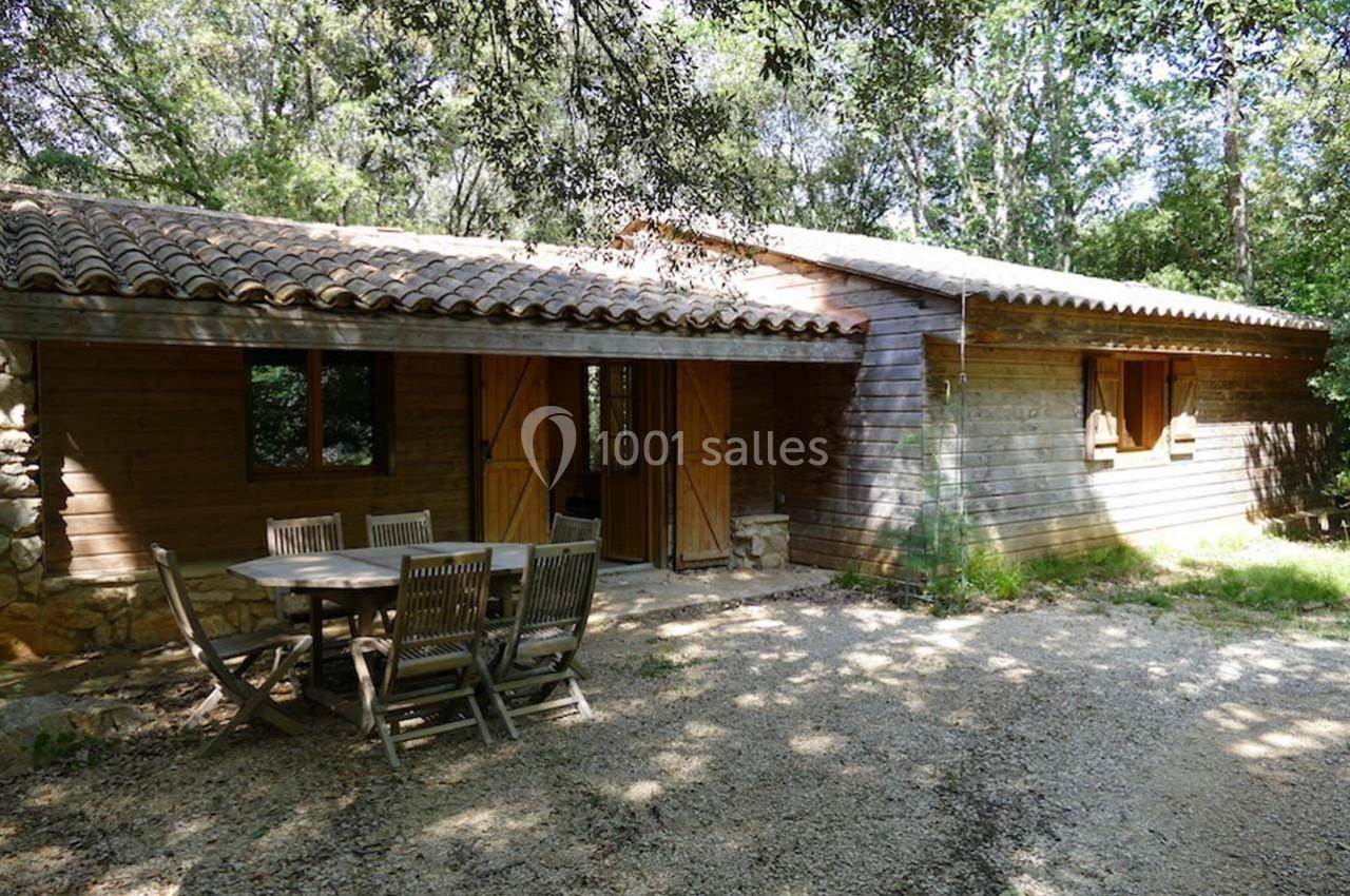 Maison en bois avec toit en tuiles, entourée d'arbres, avec une table et des chaises en bois sur une terrasse en gravier.