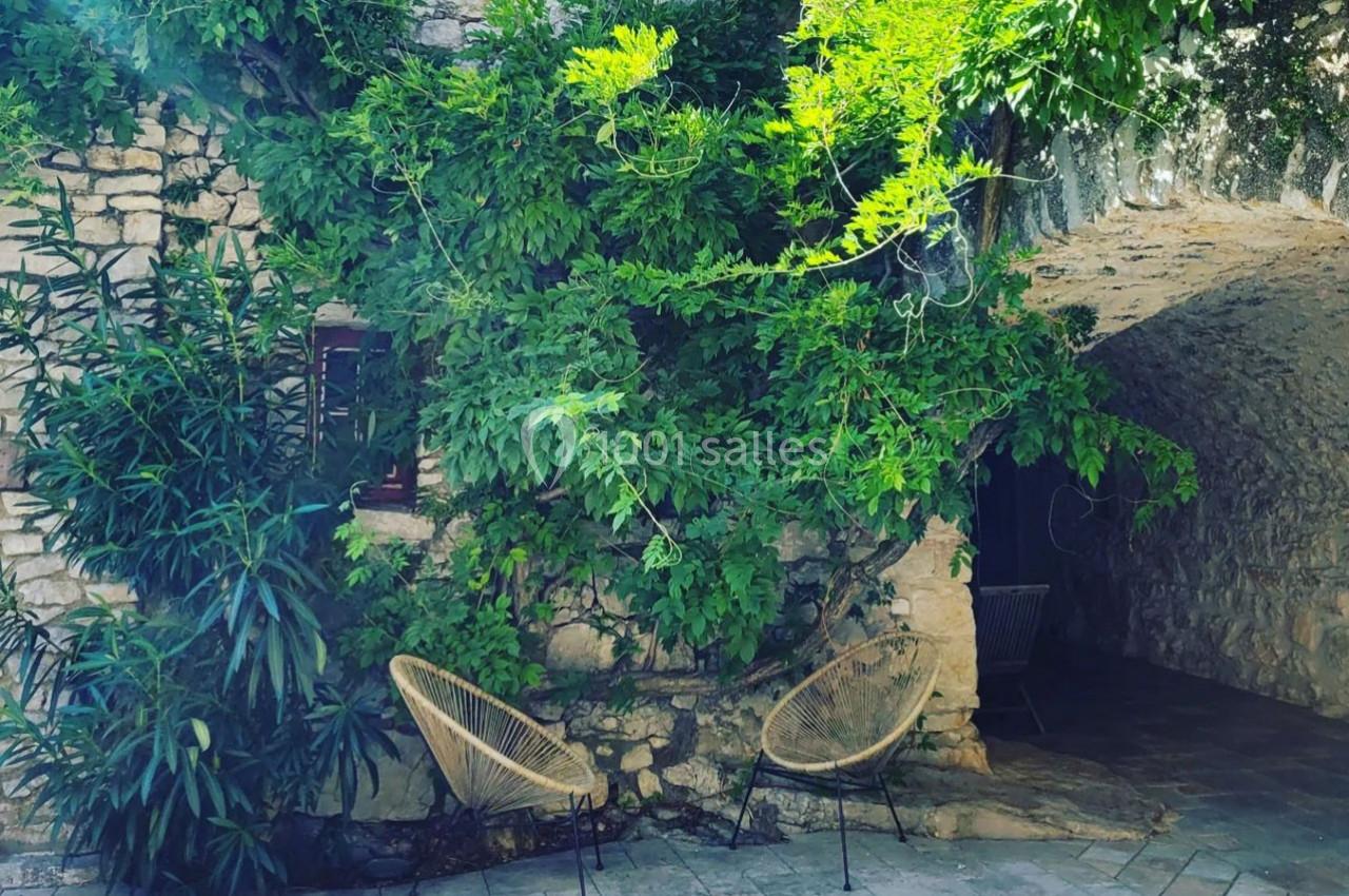 Façade en pierre recouverte de végétation avec deux fauteuils en osier sous une arche ombragée.