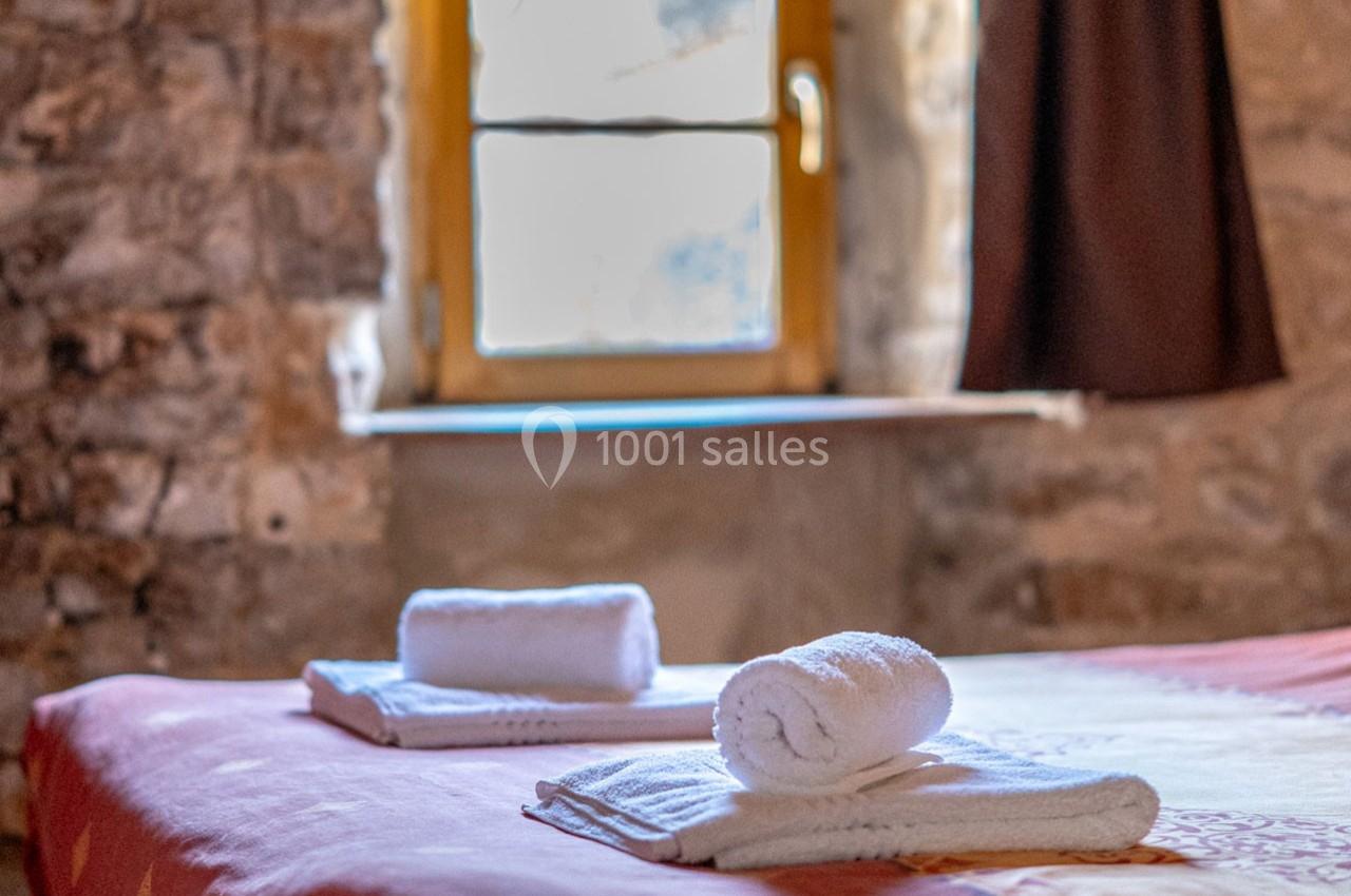 Serviettes blanches roulées posées sur un lit dans une chambre en pierre avec une fenêtre éclairée en arrière-plan.