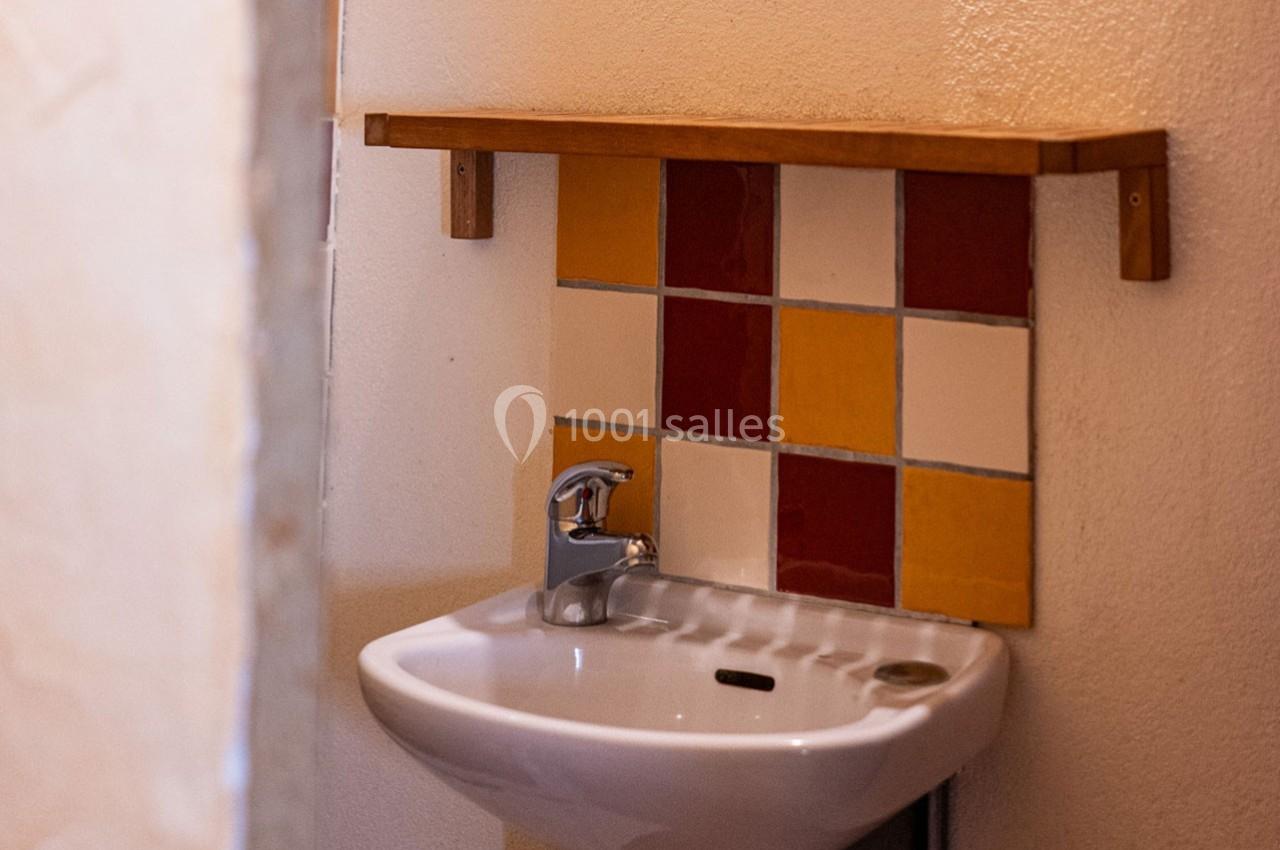 Lavabo blanc avec robinet, placé sous une étagère en bois, devant un mur décoré de carreaux colorés.