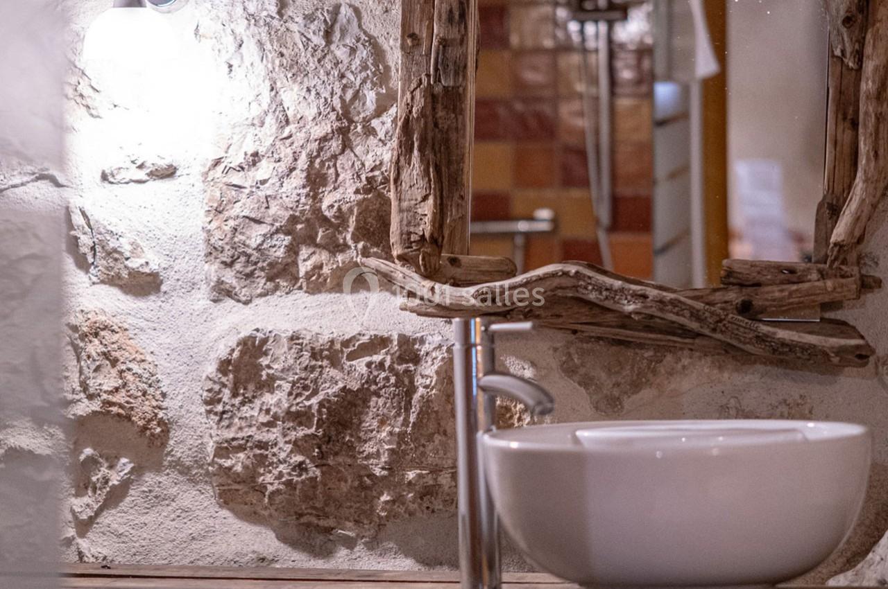 Lavabo blanc avec robinet moderne devant un miroir encadré de bois brut, sur un mur en pierre apparente.