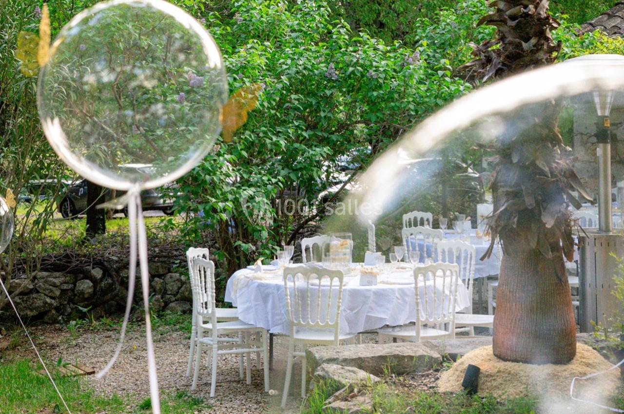 Tables dressées avec nappes blanches dans un jardin verdoyant, entourées de chaises blanches et de végétation.