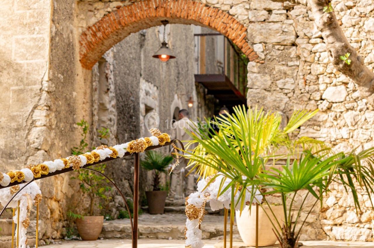 Passage en pierre avec arche en briques, décoré de fleurs blanches et dorées, entouré de plantes vertes.