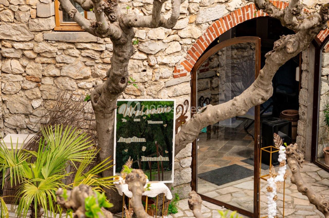 Entrée d'un bâtiment en pierre avec une porte vitrée, un arbre taillé et un panneau de bienvenue décoratif.